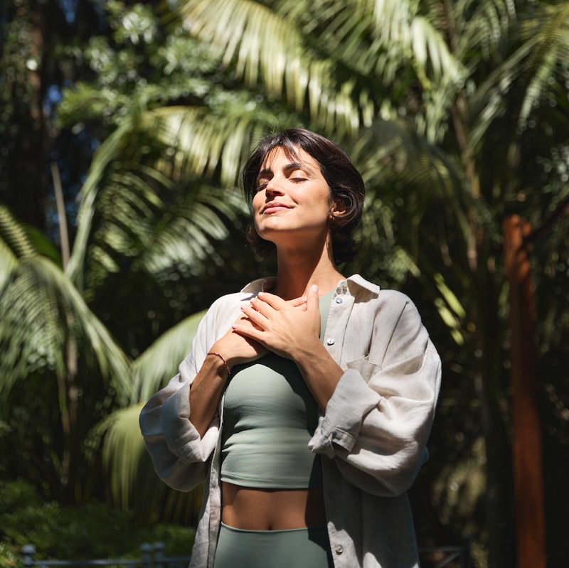 Happy calm young Hispanic woman holding hands on chest meditating doing yoga breathing exercises with eyes closed feeling gratitude, mental balance standing in green nature tropical park. Vertical.